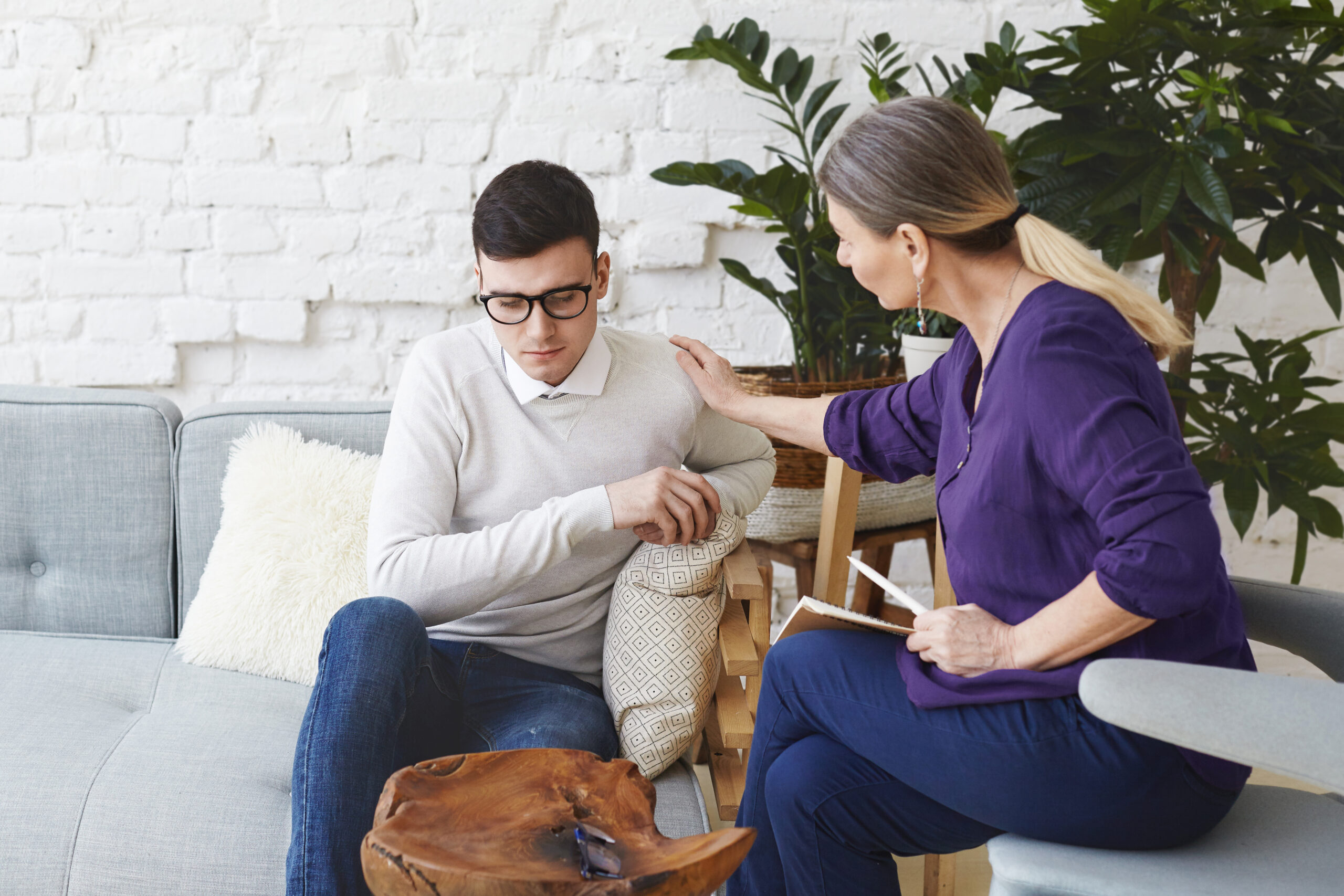 candid shot of casually dressed professional woman psychotherapist in her fifties touching her young male patient by shoulder while having counseling session, expressing sympathy and support